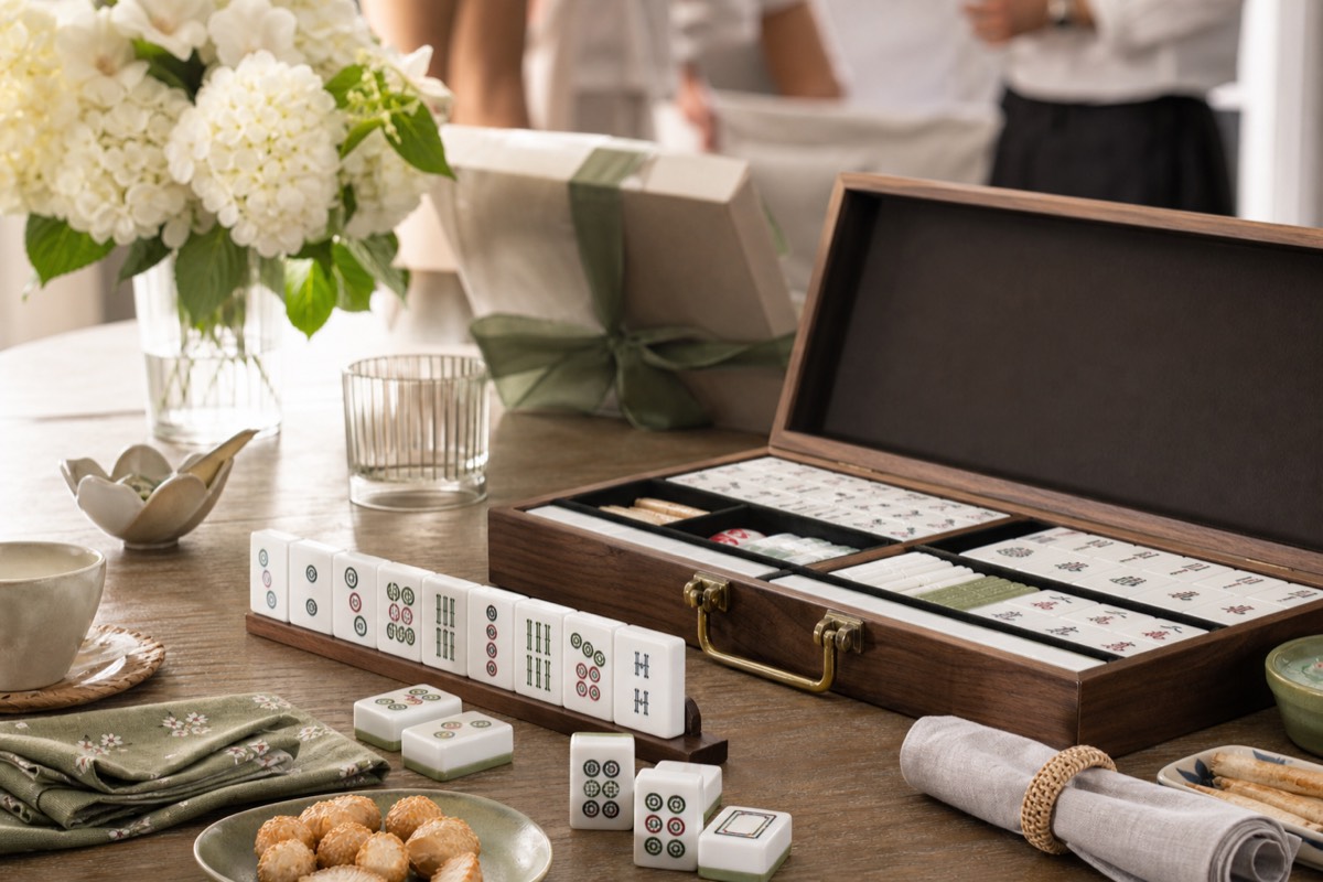 A giftable mahjong set in a wooden case arranged on a home table with flowers, tea, and snacks